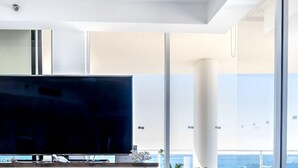 A modern living room with a large flat-screen TV, white cabinetry, a round grey ottoman, and a view of the ocean through large windows. - Panoramic Ocean Views At Maroochydore (Maroochydore Region)