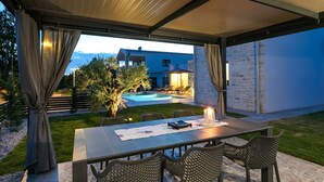 An outdoor patio with a table and chairs, a pool, and a view of a blue house. - Lovely Villa Maslina with a pool (Nova Vas)