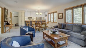 A spacious living room with a grey sofa, blue armchair, and a wooden coffee table. A kitchen with white cabinets and a dining area with wooden chairs is visible in the background. - Newly Remodeled Spacious 1 Bedroom 1 Condo by RedAwning (Avon)