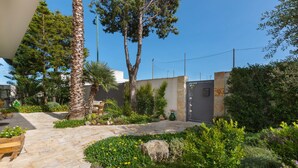 Ein Gartenpfad führt zu einem Gebäude mit einer grauen Tür, umgeben von verschiedenen Pflanzen und Bäumen.