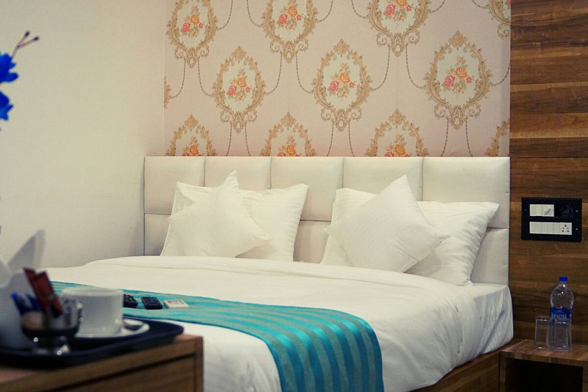 Habitación de hotel con cama, almohadas blancas, colcha azul y blanca a rayas, un florero con flores azules, mesita de noche con una botella de agua y una pared con diseño floral.