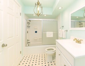 A bathroom with a white toilet, a shower with a glass door, a white sink with a gold faucet, and a chandelier. - ~ Sunrise Sands ~ Oceanfront Escape! (Myrtle Beach)