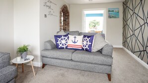 Un soggiorno con un divano grigio, cuscini a tema nautico, un piccolo tavolino con una pianta e una parete decorata con un quadro di un faro.