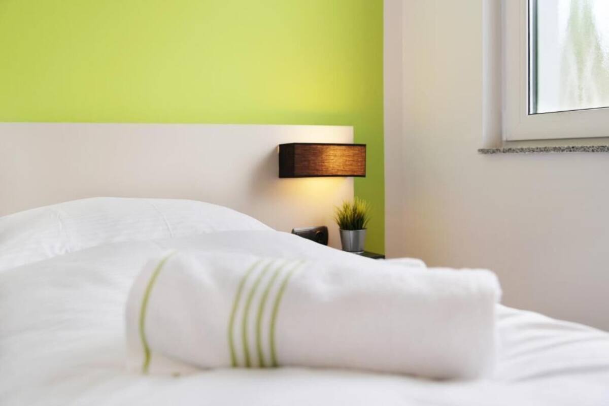 Una cama bien hecha con ropa de cama blanca y franjas verdes, una lámpara de noche y una pequeña planta en maceta sobre una mesita de noche.