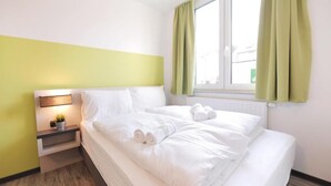 A neatly made bed with white linens and towels, a bedside table with a lamp, and a window with green curtains. - bp24 Hotel Eschweiler (Eschweiler)