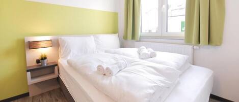 A neatly made bed with white linens and towels, a bedside table with a lamp, and a window with green curtains.