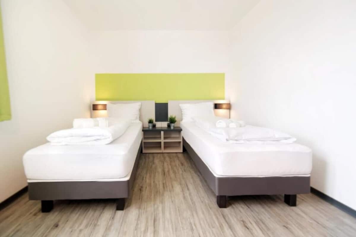 Habitación de hotel con dos camas individuales, una mesita con lámpara y una pared verde decorativa.