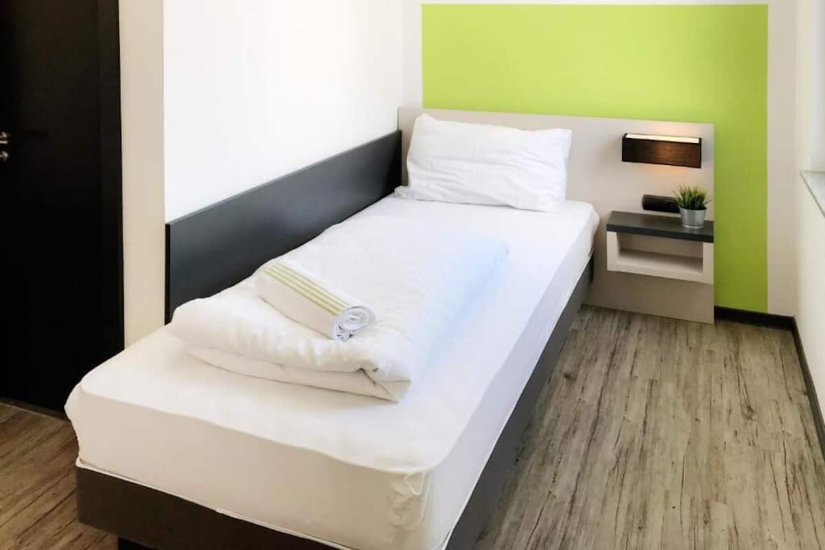 Una cama bien hecha con sábanas blancas y una toalla doblada, una mesita de noche con una lámpara y una planta pequeña, y una pared de acento verde.