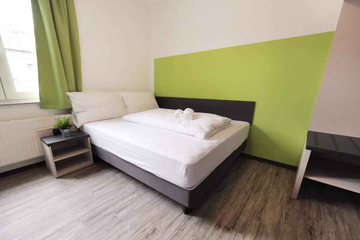 Una cama bien hecha con sábanas blancas y toallas enrolladas, una planta en la mesita de noche y una pequeña mesita de madera.