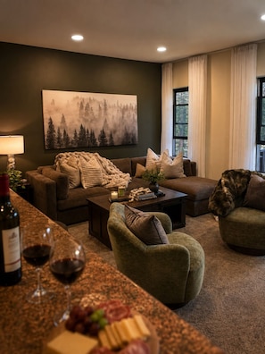 A cozy living room with a dark green wall, a large forest-themed painting, a brown sofa, a green armchair, and a coffee table with books and a candle.