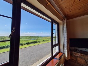 A room with a large window offering a view of a road and green landscape. - Sea View Cottage, Errislannan (Ballyconneely)