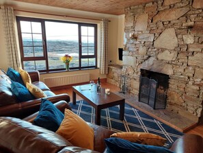 A cozy living room with a stone fireplace, leather sofa, and a view of the outdoors. - Sea View Cottage, Errislannan (Ballyconneely)