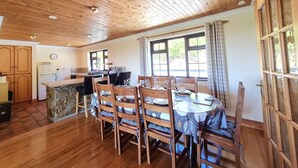 Un comedor amplio con una mesa y sillas de madera, una cocina al fondo y ventanas grandes que dejan entrar luz natural.