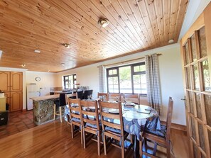 A dining area with a wooden table and chairs, a kitchen in the background, and a large window with curtains. - Sea View Cottage, Errislannan (Ballyconneely)