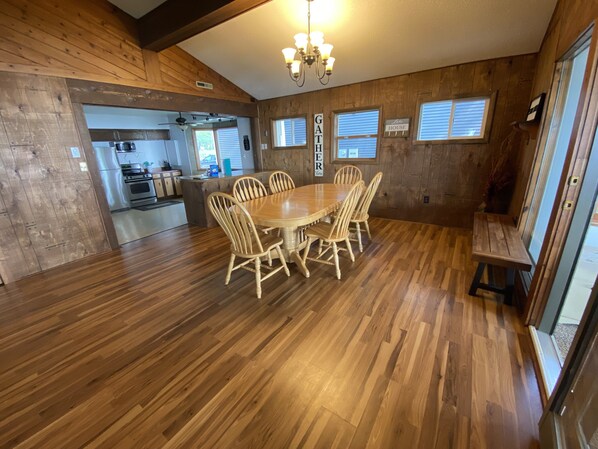 A wooden interior with a dining table surrounded by chairs, a kitchen in the background, and a sign that reads 'GATHER HERE'. - Lakefront home with 3 bedrooms, 2 baths, great for groups up to 10 (Shelbyville)