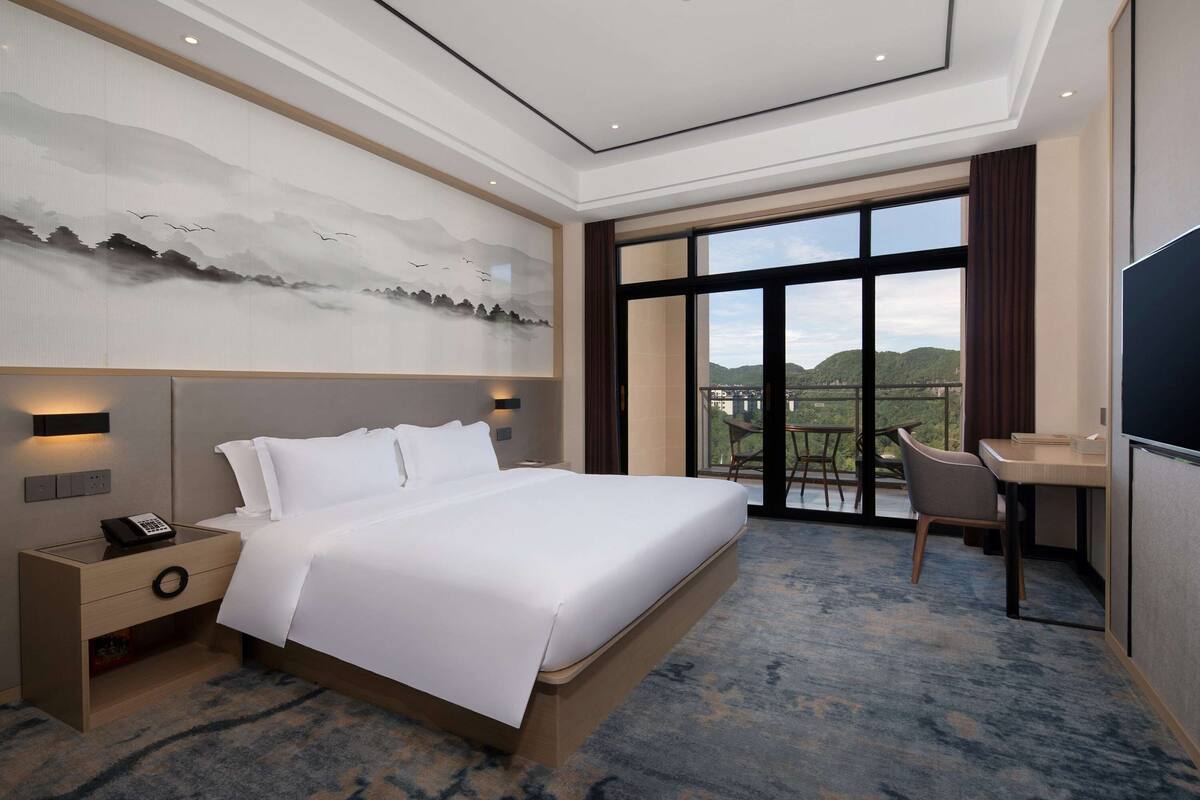 A hotel room with a large bed, a desk, a chair, a nightstand with a phone, and a large window with a view.
