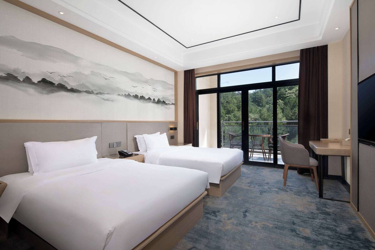 A hotel room with two beds, a desk, a chair, and a large window offering a view of the outdoors.