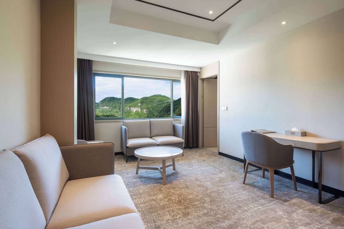 A modern hotel room with a sofa, a small round table, a desk, and a view of green hills through large windows.