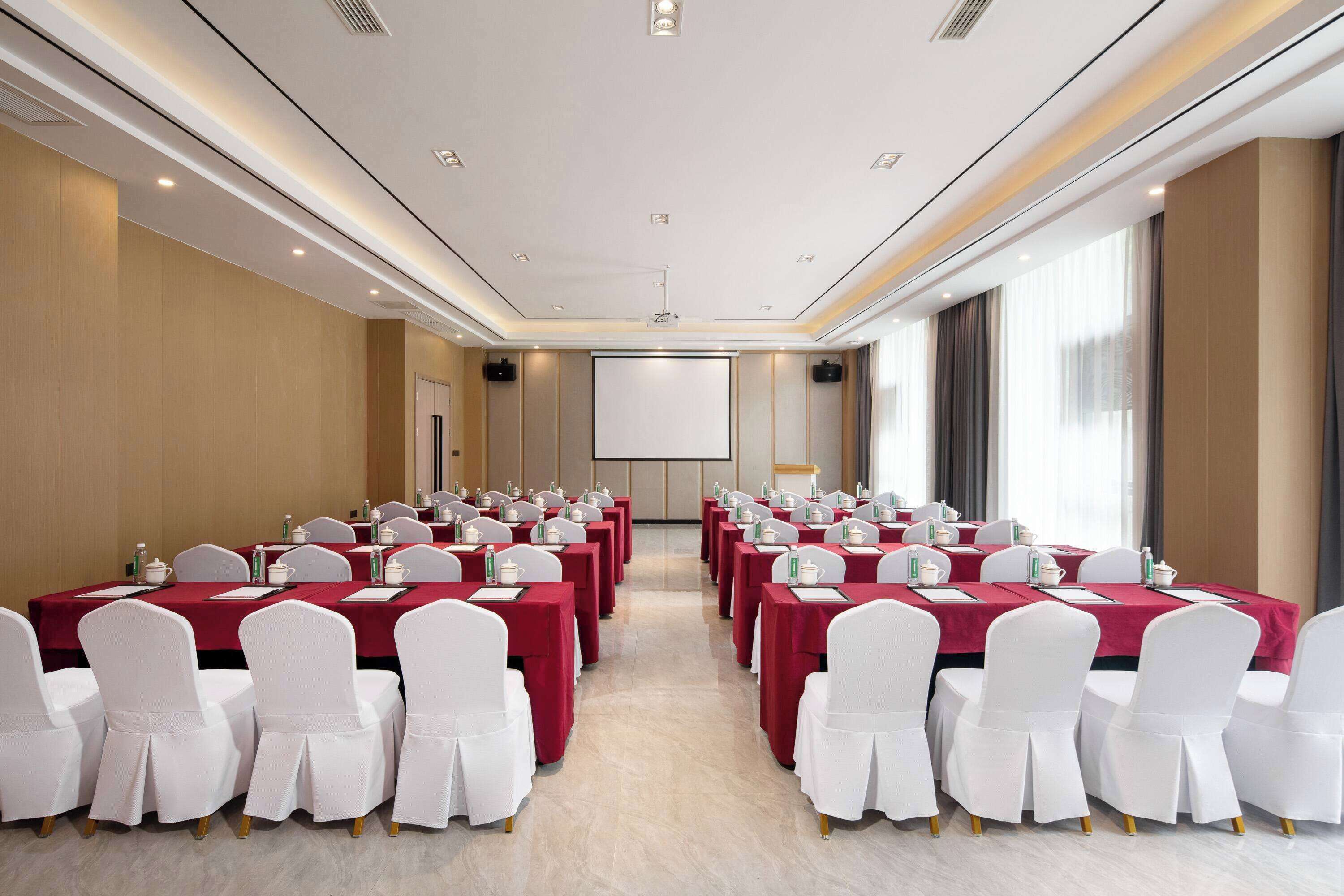 A conference room with rows of tables and chairs, a projector screen, and a large window with curtains.