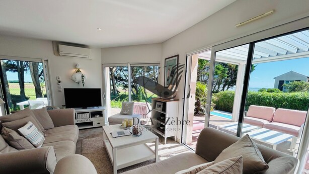 Una sala moderna con un sofá color beige, una mesa de centro y un televisor. Hay una puerta corrediza de vidrio que da a un área exterior con piscina y vegetación.