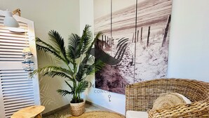 Una habitación con una silla de mimbre, un taburete de madera, una planta en maceta, una lámpara de pared y un cuadro abstracto de gran tamaño.