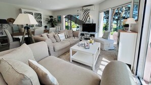 Una sala de estar moderna con un sofá color beige, una mesa de centro y un televisor. Se ve una escalera que sube a un nivel superior, y ventanas amplias que dejan entrar luz natural.