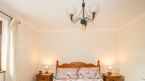 A bedroom with a wooden bed, floral bedding, two bedside tables with lamps, a chandelier, and a window with curtains. - Penrallt Fawr (Caernarfon)