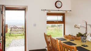 A dining area with a table set for two, a wooden chair, a clock on the wall, and a view of the outdoors through a window. - Penrallt Fawr (Caernarfon)
