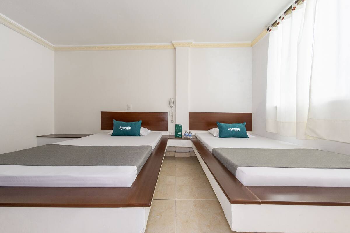 A hotel room with two beds, each with a wooden headboard and a teal pillow. The room has white walls and a tiled floor.