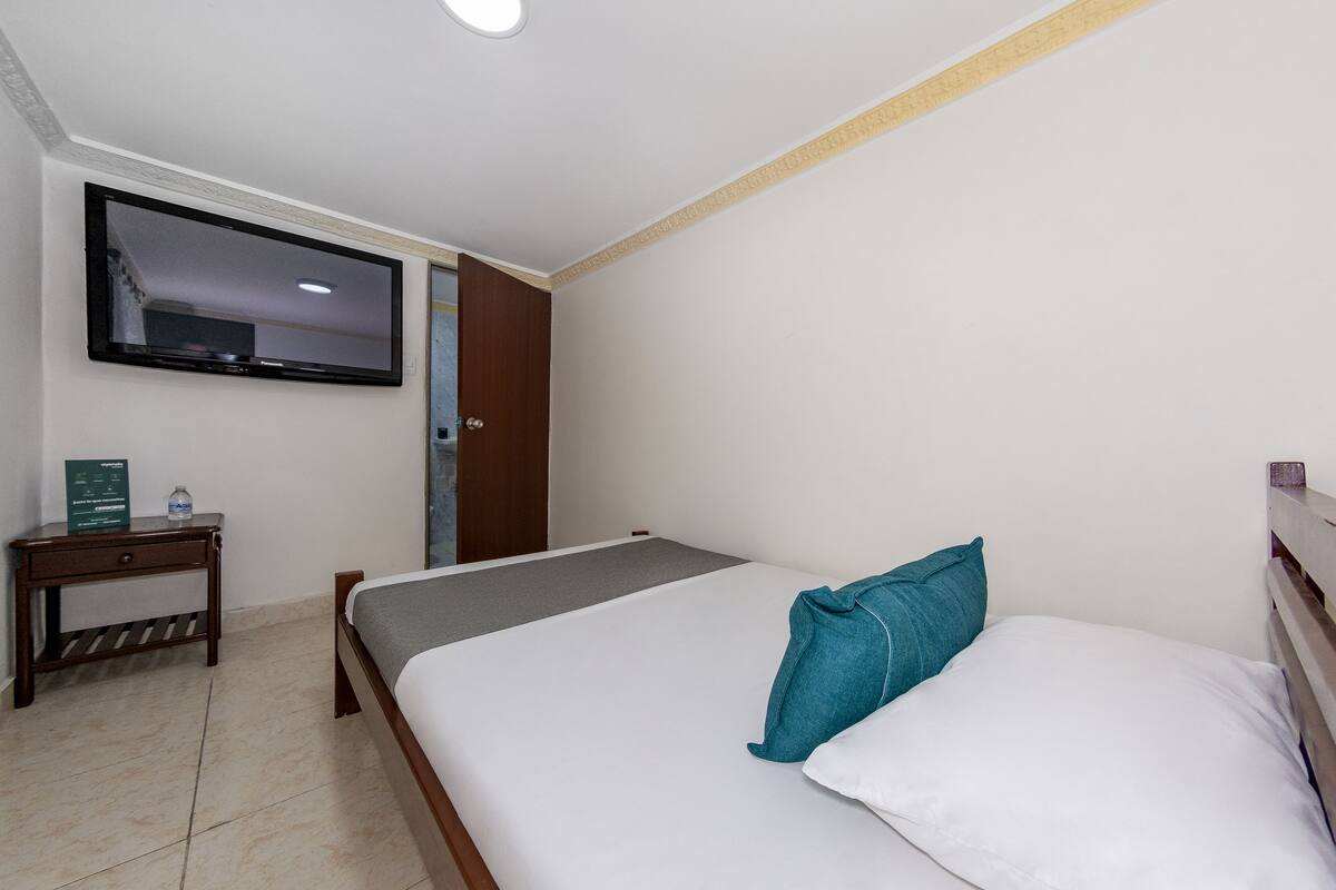 A hotel room with a bed, a TV mounted on the wall, a small table with a water bottle, and a door.