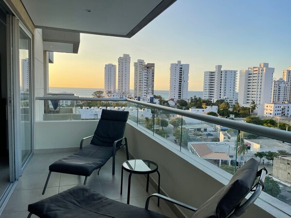 Un balcon avec deux fauteuils et une petite table, offrant une vue sur le paysage urbain avec ses grands immeubles et la mer.