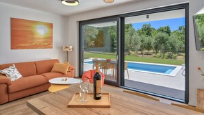 Ein modernes Wohnzimmer mit einem großen Glastür zum Poolbereich, einem Sofa, einer Weinflasche und einem Gemälde eines Sonnenuntergangs.