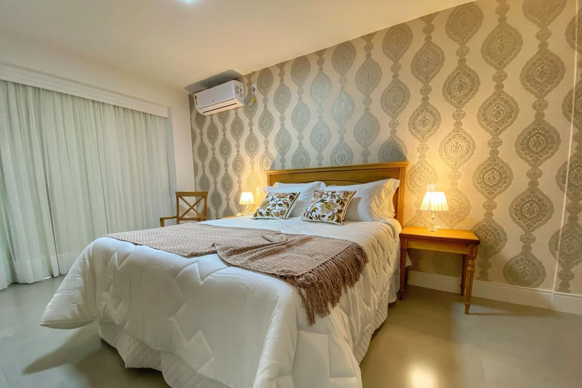 A hotel room with a large bed, two bedside tables with lamps, a wall-mounted air conditioner, and patterned wallpaper.
