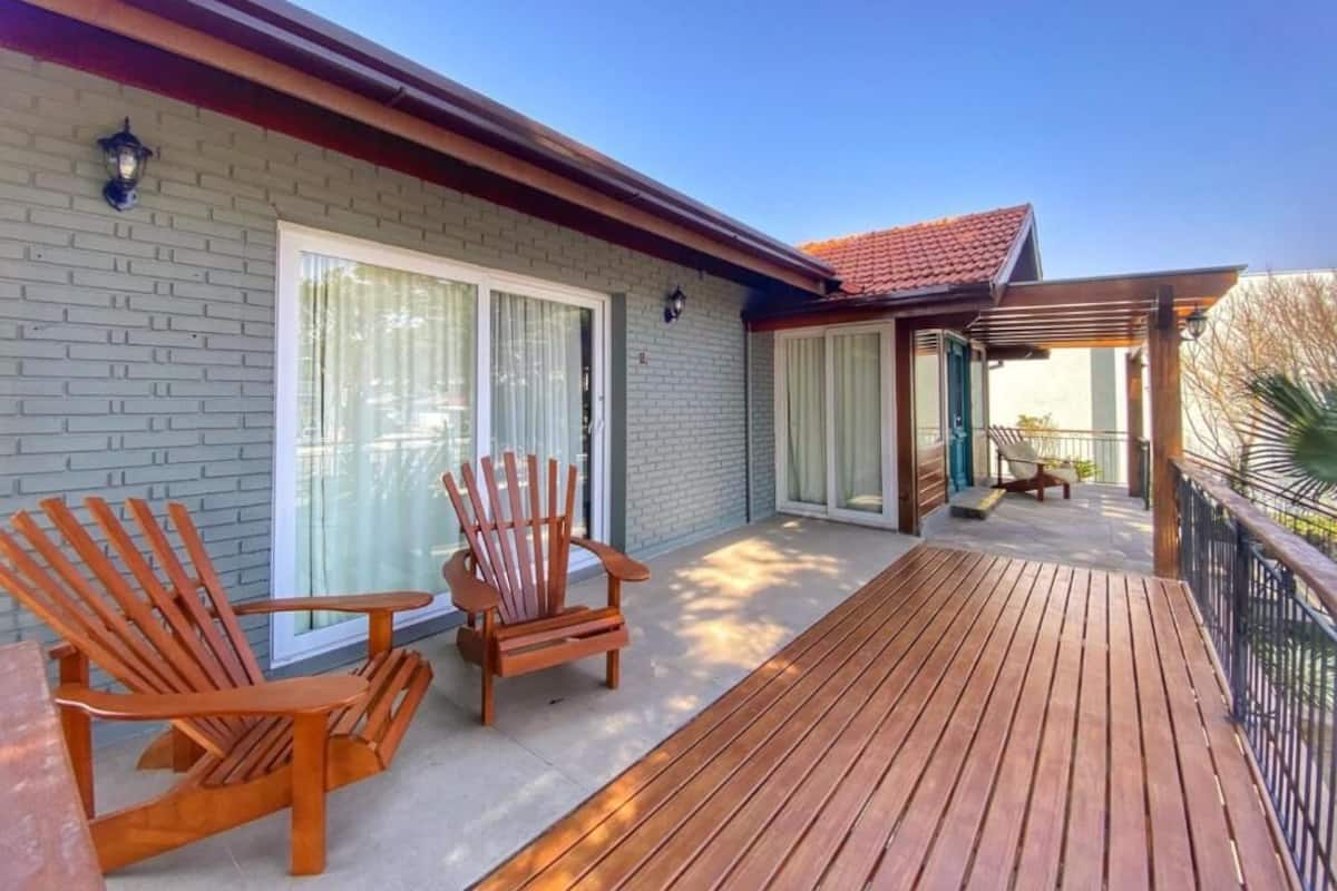 A wooden deck with two Adirondack chairs, a covered patio area, and a brick building with a red roof.