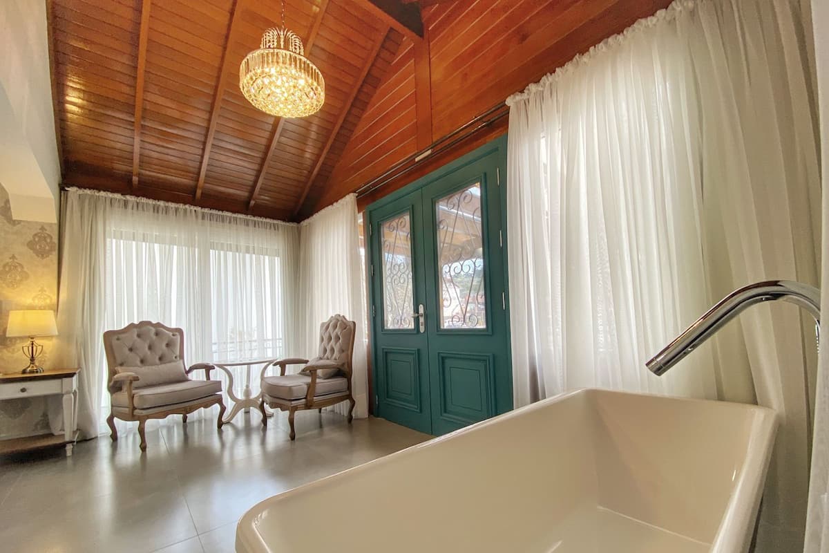 A spacious room with a large bathtub, two upholstered chairs, a wooden ceiling, and a green door.