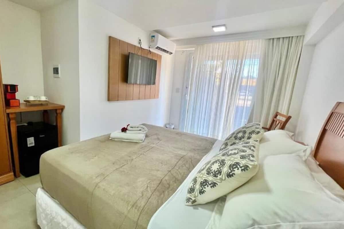 A hotel room with a bed, a TV mounted on the wall, a window with curtains, and a wooden wardrobe.
