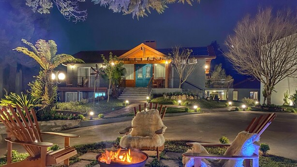 A well-lit outdoor seating area with a fire pit, surrounded by wooden chairs and a small table, in front of a house with a porch and steps leading to the entrance. - Pousada Ville Vie (Gramado)