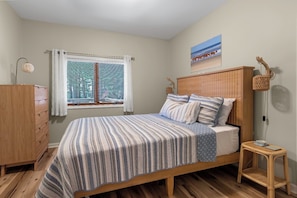 A bedroom with a bed, wooden dresser, window with blinds, and a wall-mounted lamp.
