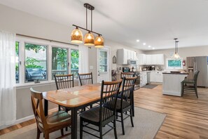 A modern kitchen with white cabinets, a dining area with a wooden table and chairs, and a view of the outdoors through large windows. - Secluded 4BR Cottage Near Lake Michigan—Decks, Fire Pit & Pet‑Friendly (South Haven)