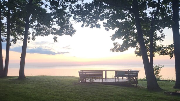 A grassy area with a bench and a table, surrounded by trees, overlooking a calm body of water. - Secluded 4BR Cottage Near Lake Michigan—Decks, Fire Pit & Pet‑Friendly (South Haven)