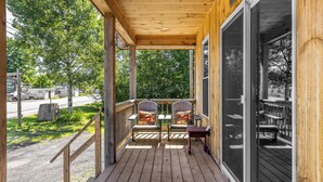 Un portico in legno con due sedie e un tavolo, affacciato su una strada e del verde.