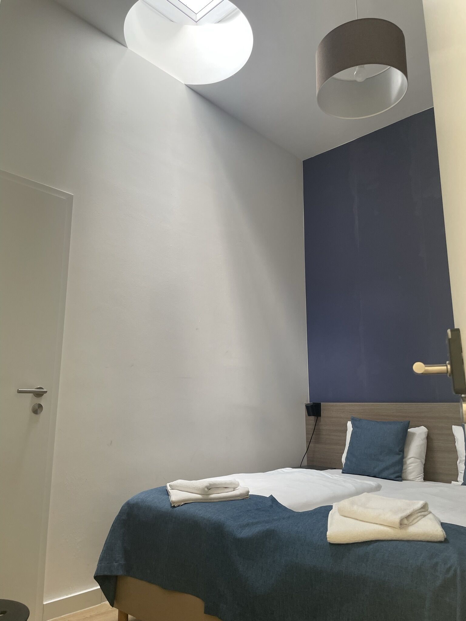 A hotel room with a bed, a bedside table, a wall-mounted lamp, and a door.
