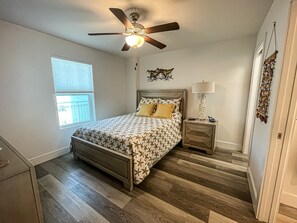 Une chambre à coucher comprenant un lit, une table de chevet, un ventilateur de plafond, une fenêtre et un élément décoratif mural.