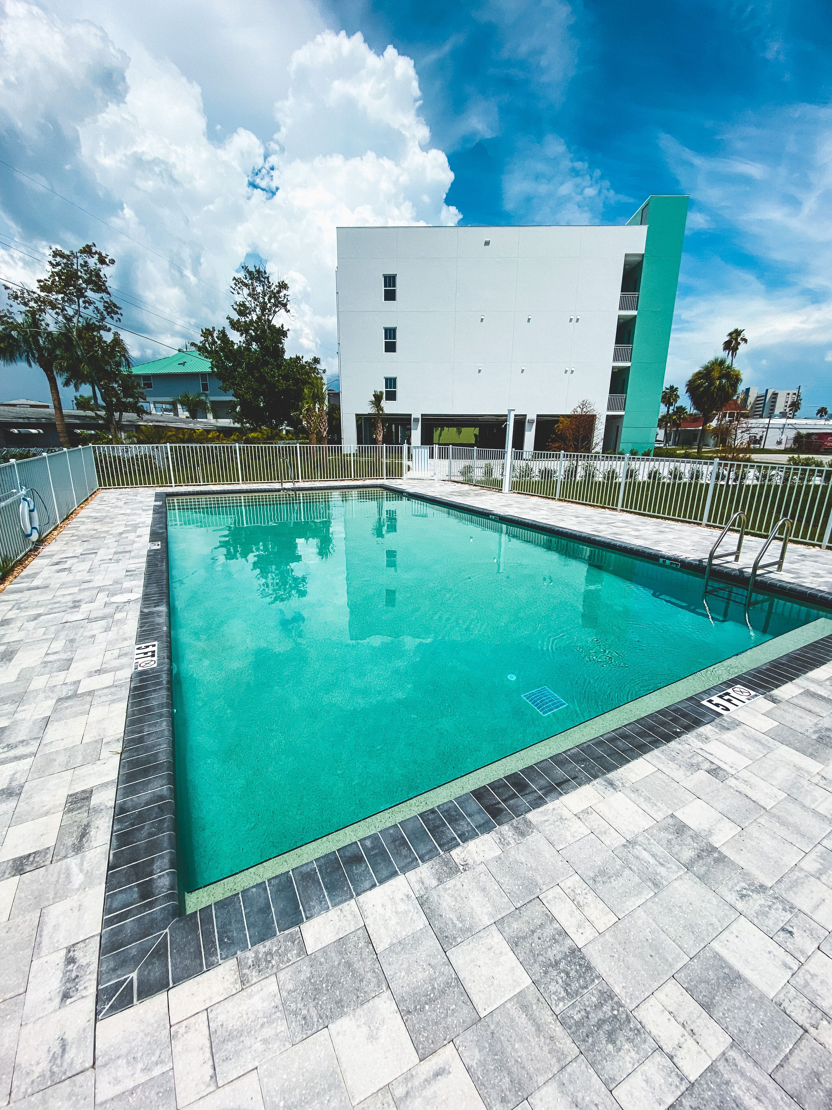 A rectangular swimming pool with a tiled deck, surrounded by a white fence, and a modern building with a green accent in the background.