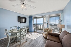 A room with a dining table, chairs, a sofa, a ceiling fan, a TV mounted on the wall, and a view of the ocean through a sliding glass door. - Sea Cabin 122 - 1 Bedroom Ocean Front Condo (North Myrtle Beach)