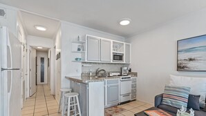 Une cuisine de taille réduite avec des meubles blancs, un four à micro-ondes et une cuisinière. On y trouve un tabouret de bar et une table basse avec un livre et un objet décoratif. Un canapé avec des coussins à rayures est visible au fond.