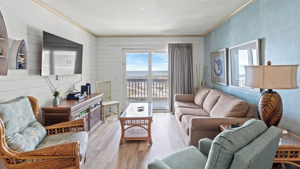 A living room with a large window, a flat-screen TV, wicker furniture, and a view of the beach. - Sea Cabin 208 - Ocean View 2 Bed/2 Bath Condo (North Myrtle Beach)
