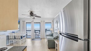 A modern kitchen with a stainless steel refrigerator, a dining area with white chairs and a table, and a balcony with a view of the ocean.