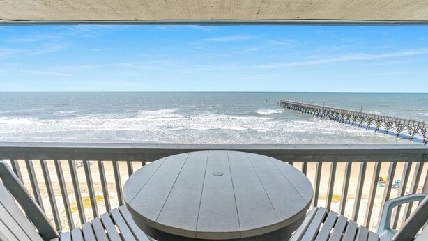 A balcony with a round table and two chairs, overlooking a beach with waves and a pier.