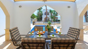 A covered outdoor dining area with a pool view, featuring a table set for breakfast with chairs and a view of the pool and buildings in the background. - Protaras Villa Elena 3 (Protaras)